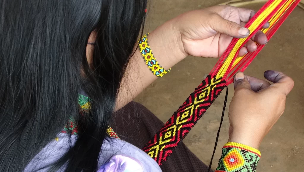 Mujer Huni Kuin tejiendo a mano una cinta con diseños kene en hilos rojos, amarillos y negros, con pulseras de mostacillas y dedos manchados de jenipapo